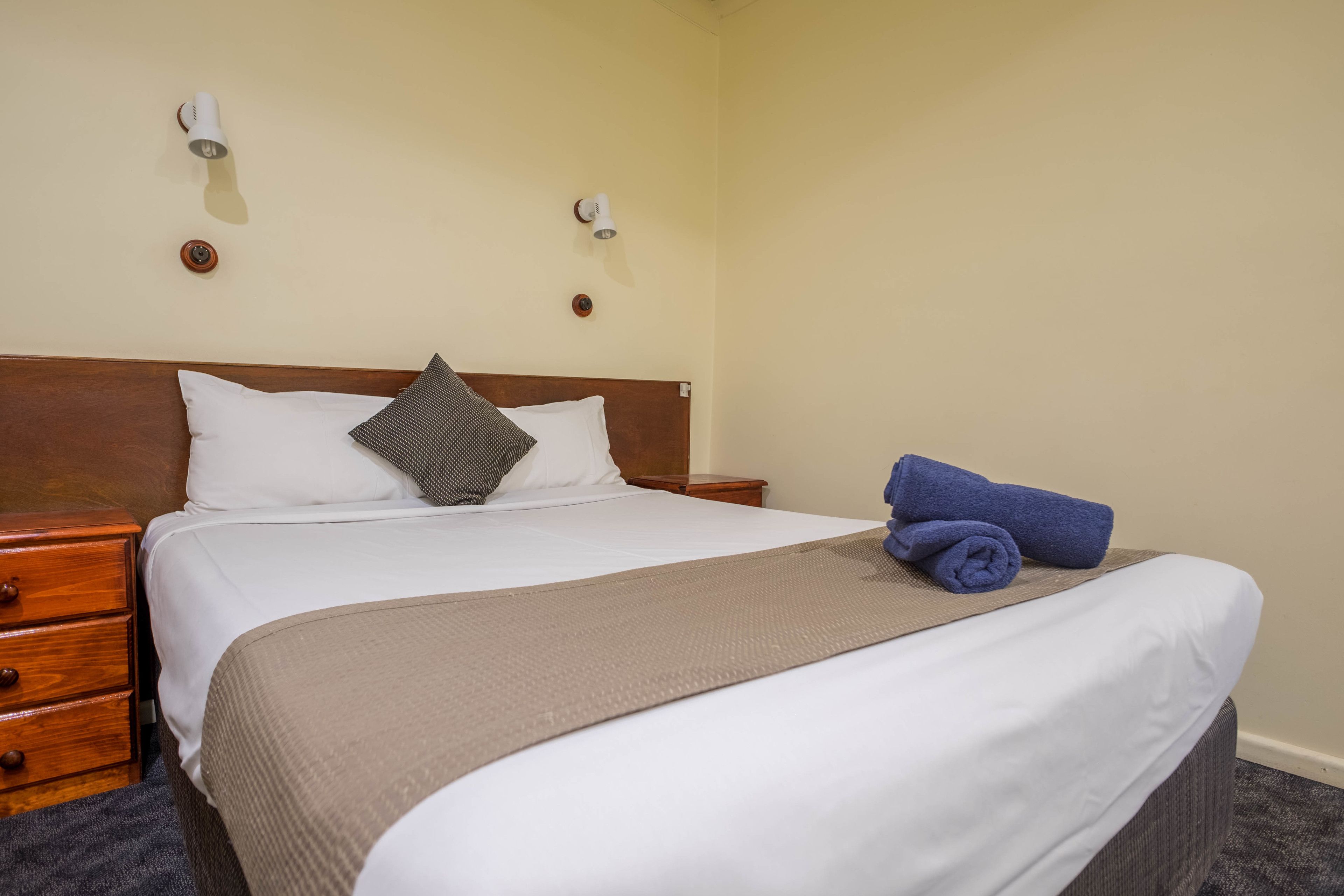 A neatly made bed with white and gray pillows, a brown throw blanket, and a blue towel on the side. There are two wall-mounted light fixtures and a wooden nightstand with a drawer.