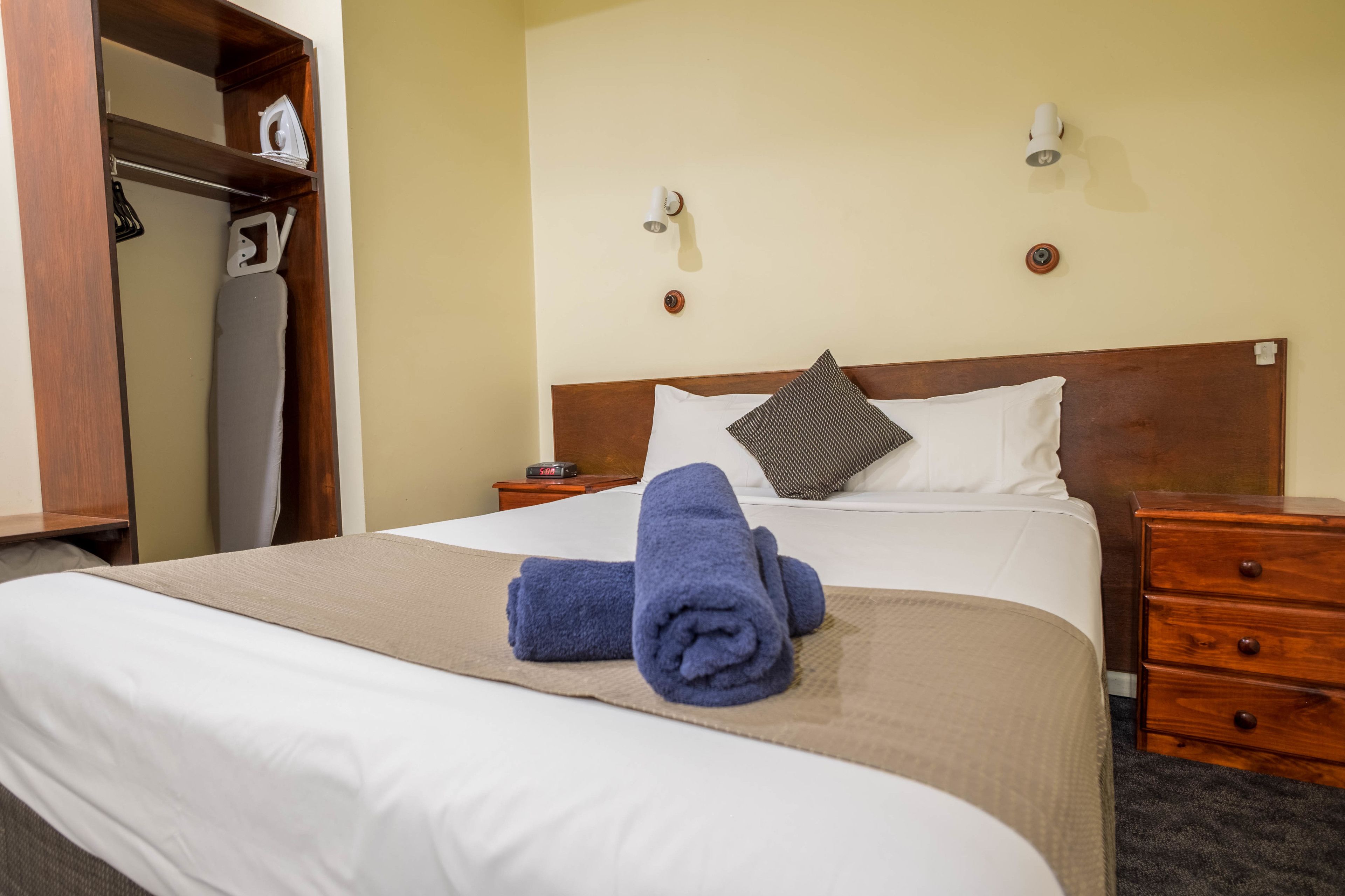 A neatly made bed with a white and beige bedspread, two pillows, and two rolled towels. There is a wooden nightstand with a lamp and a small table with an iron and ironing board.