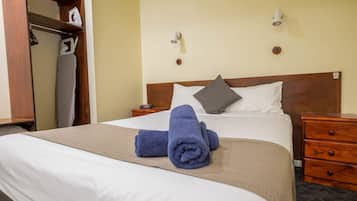 A neatly made bed with a white and beige bedspread, two pillows, and two rolled towels. There is a wooden nightstand with a lamp and a small table with an iron and ironing board.