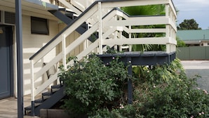 Una escalera con barandilla blanca lleva a un balcón, rodeado de follaje y un edificio con tejado verde.