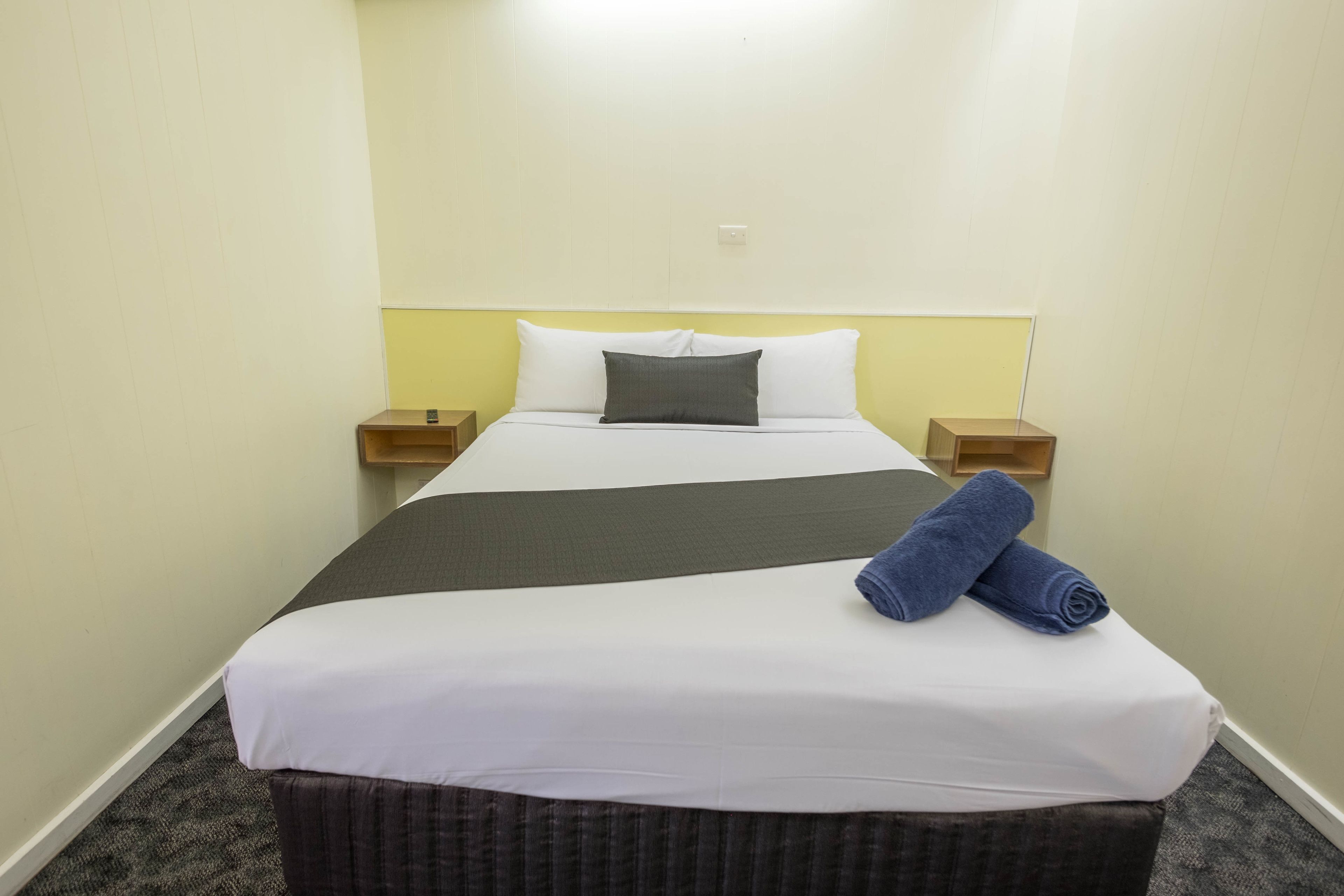 A neatly made bed with white and gray bedding, two pillows, and a folded blue towel. There are two wooden nightstands on either side of the bed.