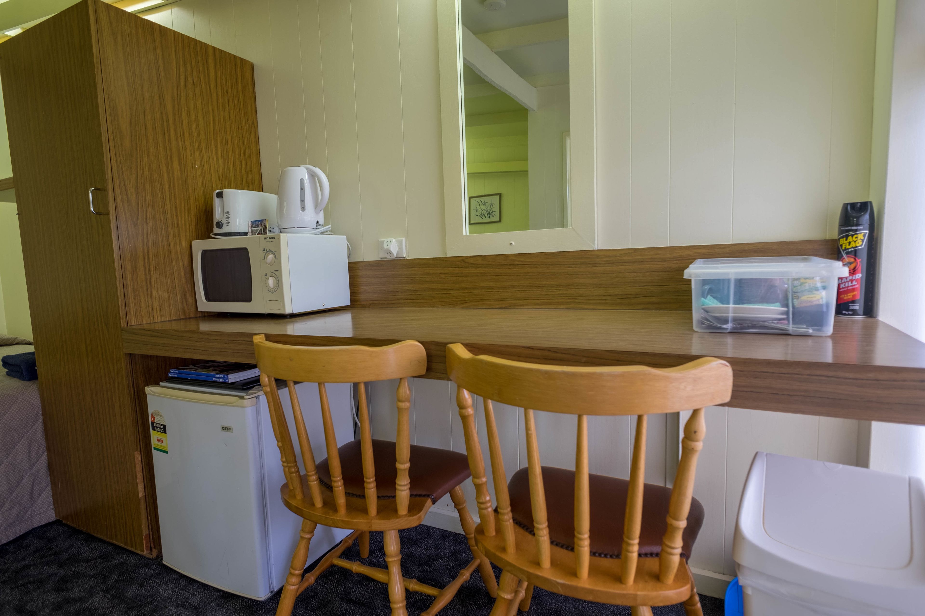 A compact kitchenette with a microwave, toaster, and small refrigerator. There are two wooden chairs and a plastic container on the counter.