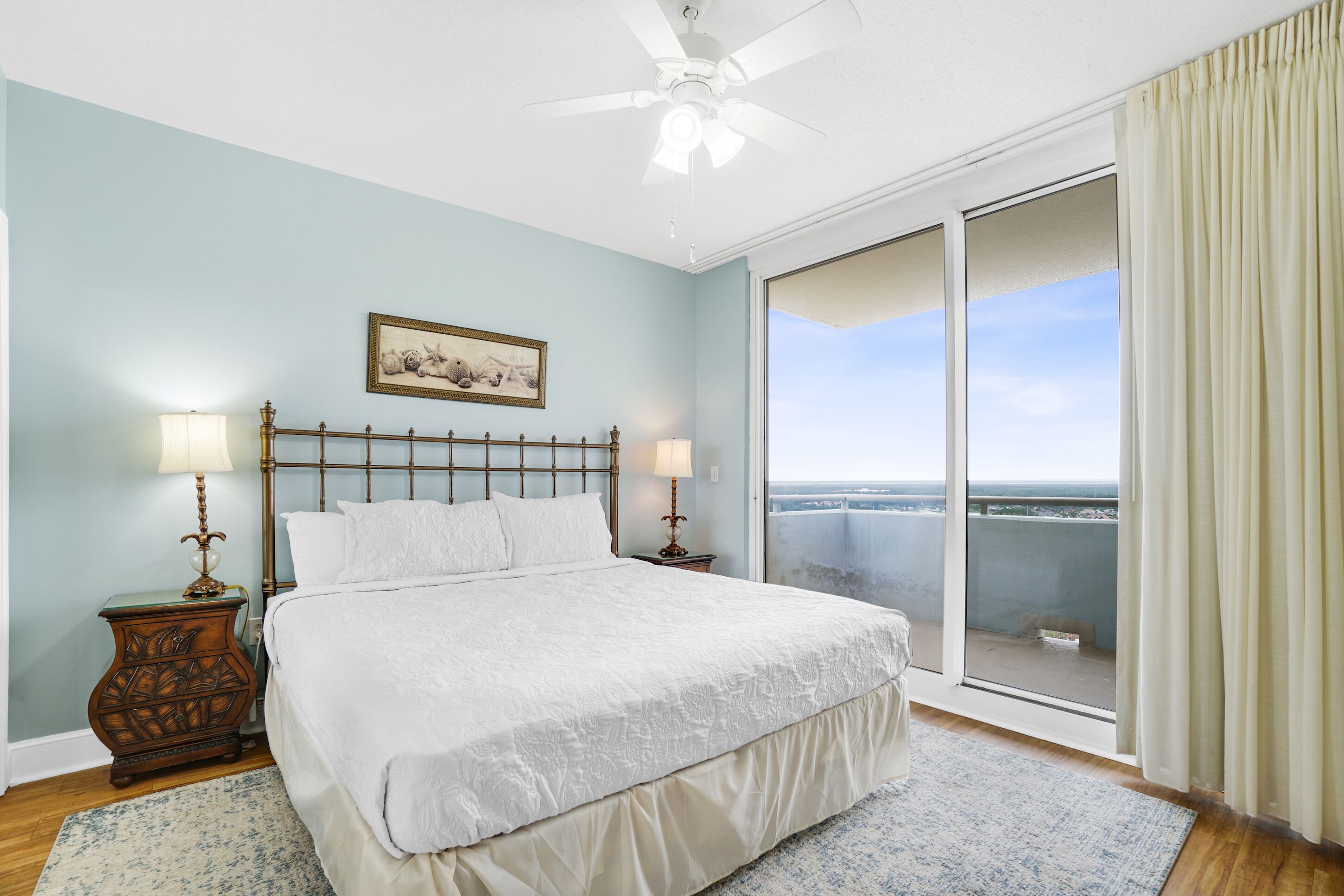 A bedroom with a large bed, two bedside tables with lamps, a ceiling fan, and a view of the city through a sliding glass door.