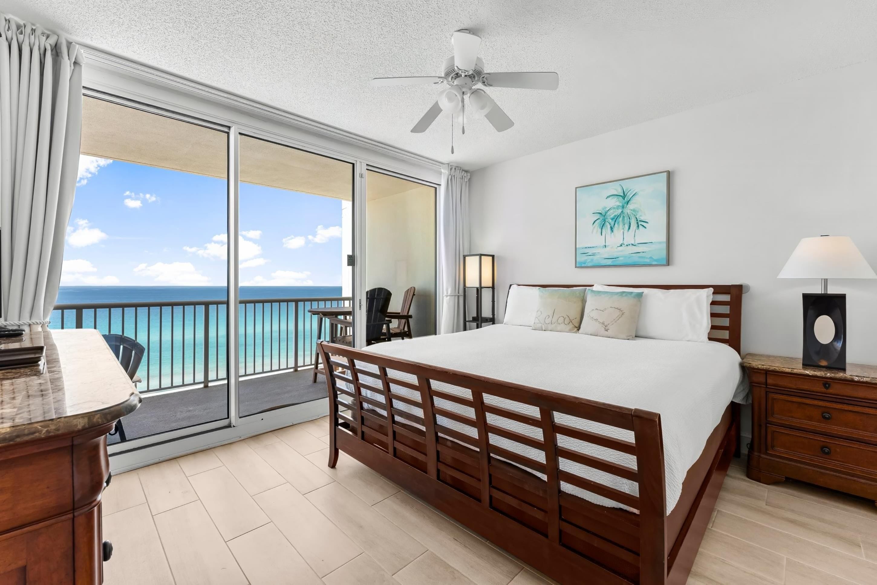 A hotel room with a large bed, a ceiling fan, a view of the ocean, a painting of palm trees, and a nightstand with a lamp.