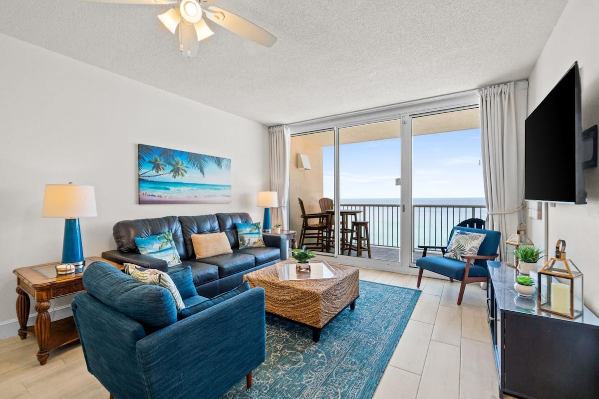 Un salon moderne avec un canapé, une table basse et des fauteuils. Un tableau représentant une scène de plage est accroché au mur, et un téléviseur y est fixé.