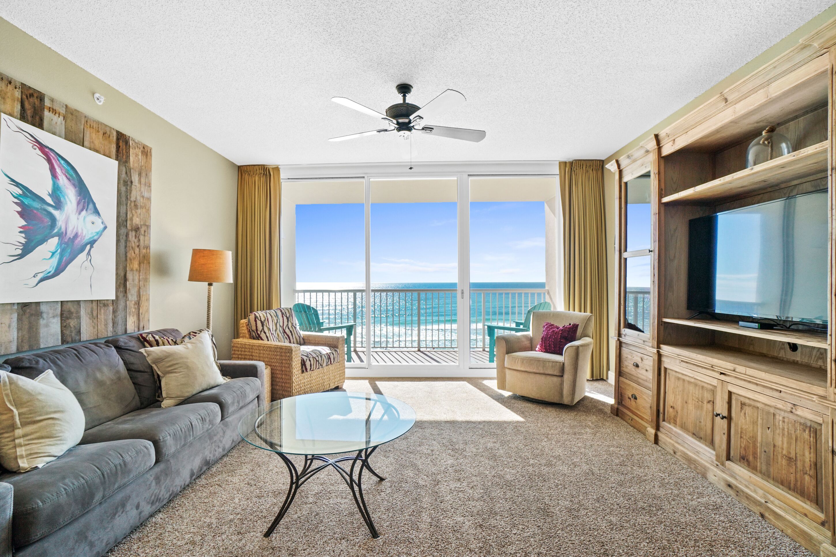 A spacious living room with a balcony, a ceiling fan, a flat-screen TV, and a large painting.