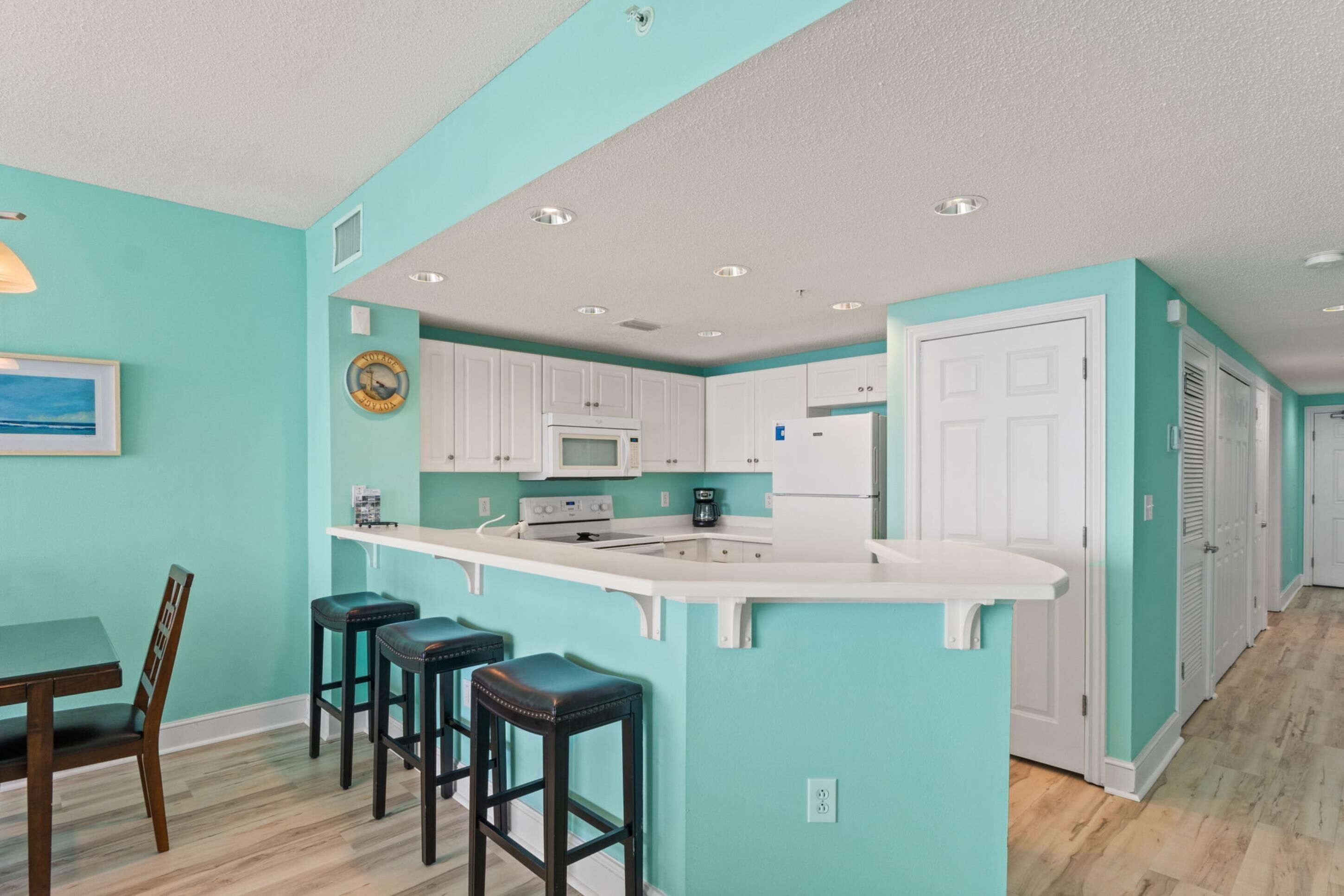 A kitchen with white cabinets, a white countertop, and a built-in oven. There are three bar stools and a wooden dining table with chairs.