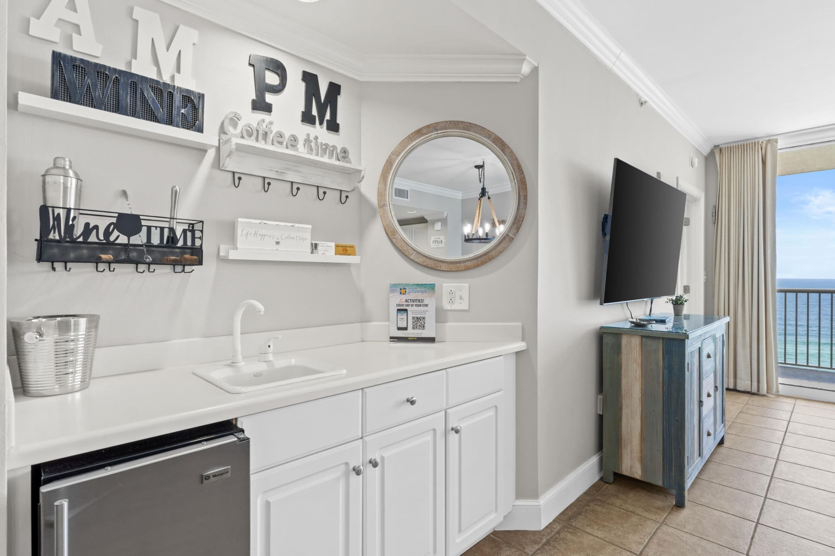 Una cocina moderna con armarios blancos, un lavaplatos empotrado y un refrigerador. Hay letras decorativas en la pared y un espejo redondo sobre el lavaplatos.