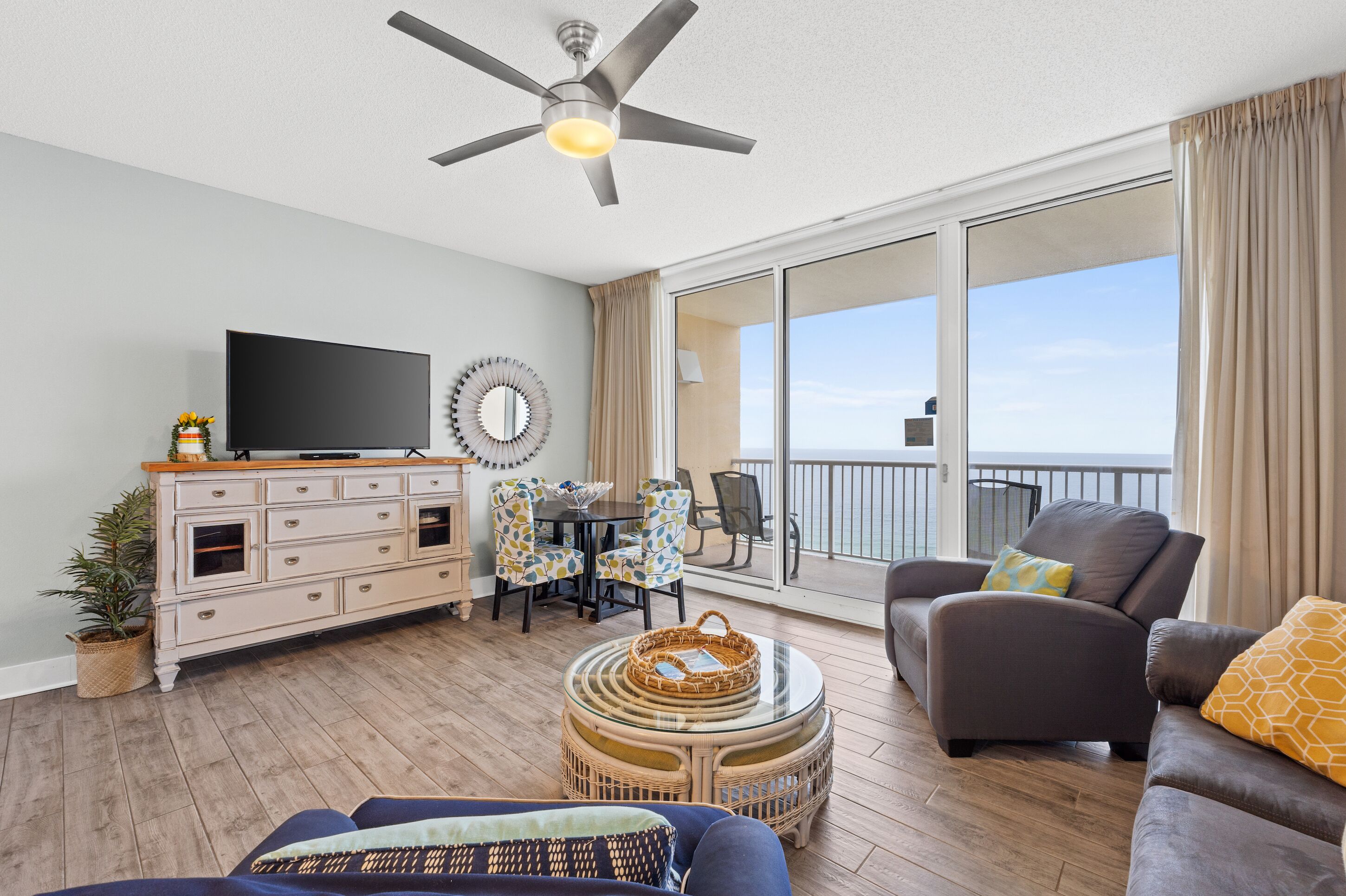 A spacious balcony with a ceiling fan, a flat-screen TV, a dining area with chairs, and a sofa with cushions.