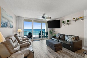 A living room with a sofa, ottoman, and chairs, a ceiling fan, a TV mounted on the wall, and a balcony with a view of the sea.