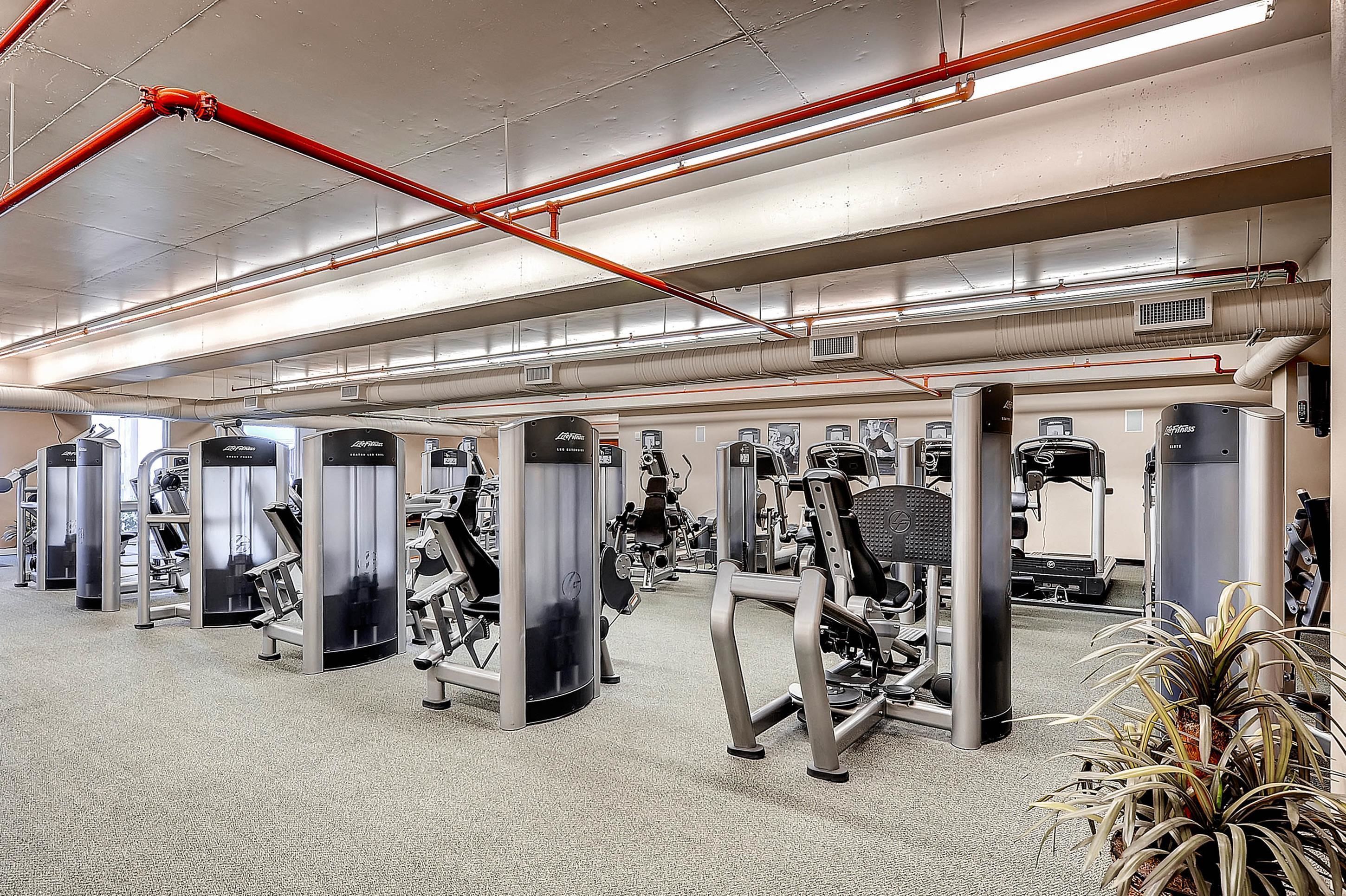 A gym with various exercise machines, red pipes on the ceiling, and air conditioning vents.