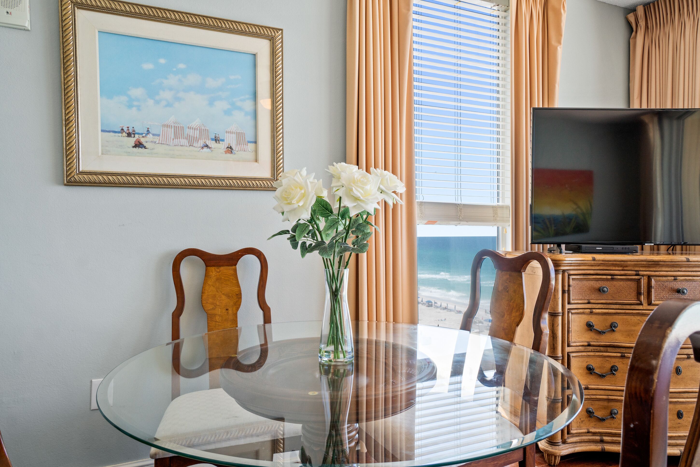 A dining area with a glass table, chairs, a vase with flowers, a framed picture on the wall, a television, and a window with curtains.