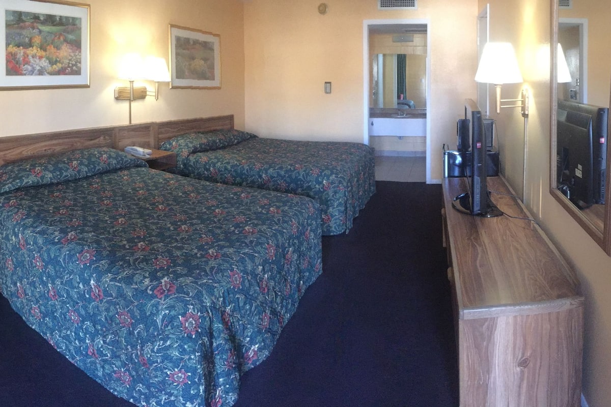 A hotel room with two beds, a small table, a chair, a desk, a mirror, and a television.