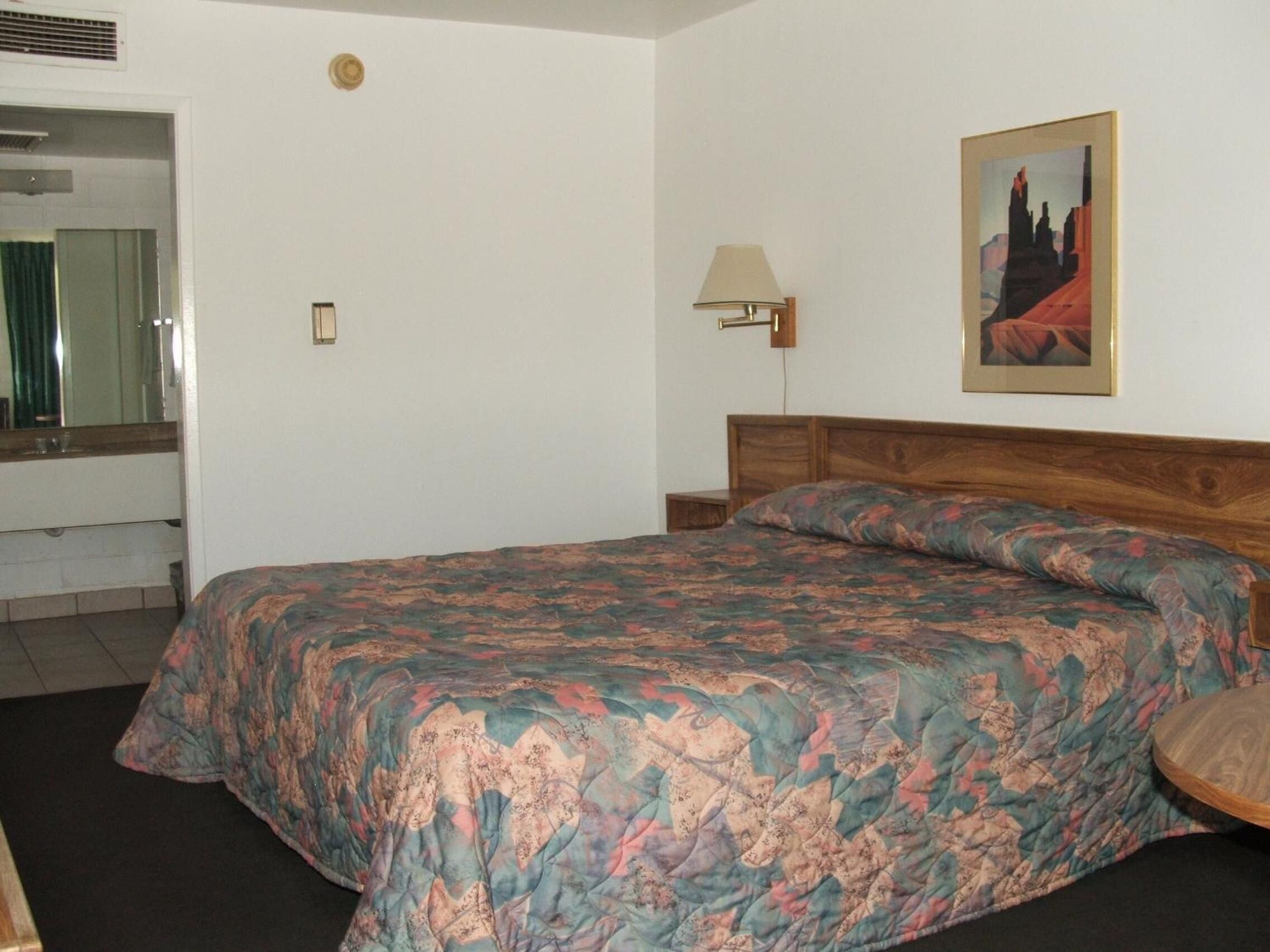 A hotel room with a large bed, a small bedside table, a lamp, a mirror, and a painting on the wall.