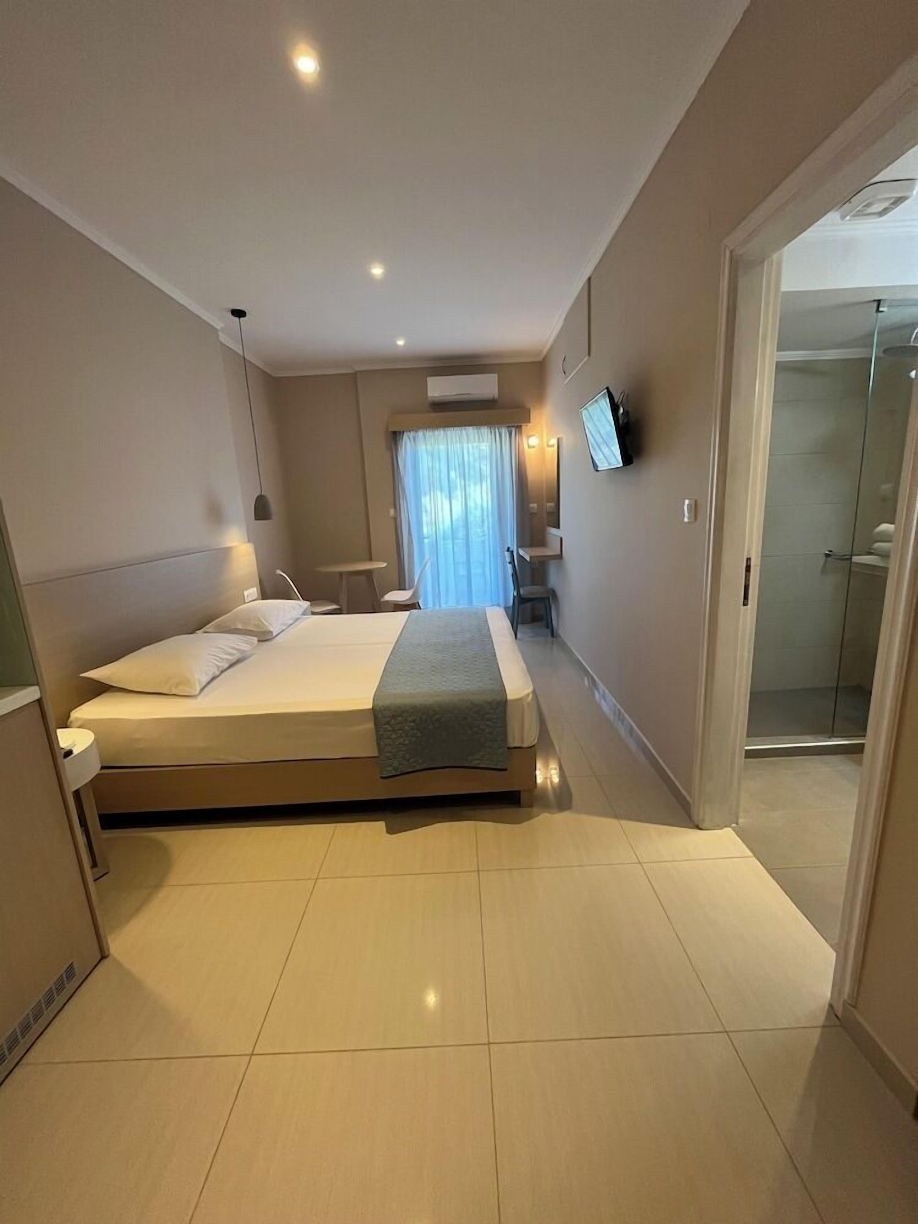A modern hotel room with a large bed, a desk, a chair, a TV, and a shower area.