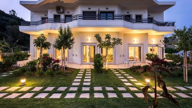 A modern two-story house with a balcony, surrounded by a well-maintained garden with stepping stones leading to the entrance.