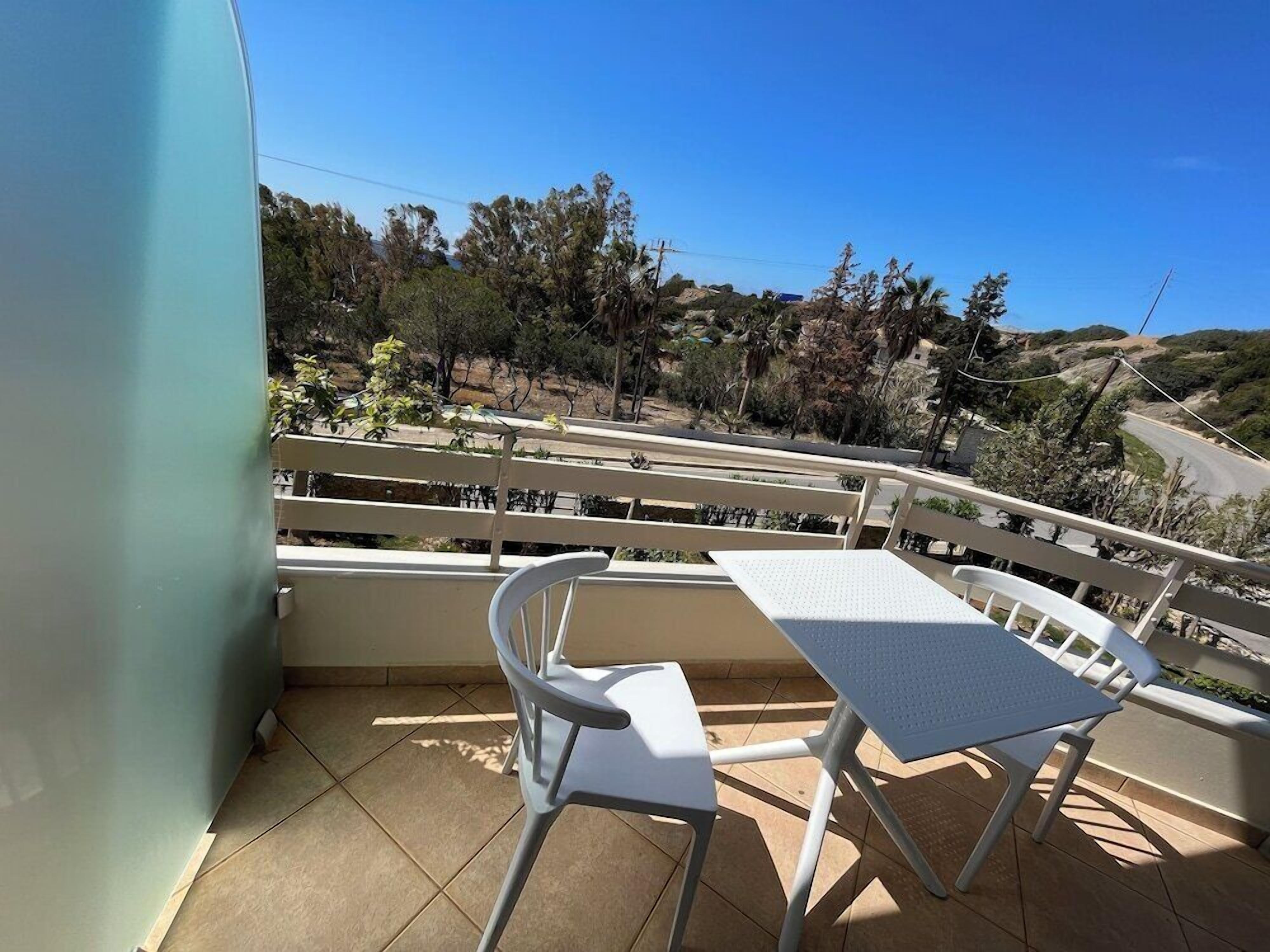 A balcony with a table and chairs, overlooking a hilly landscape with trees and a road.