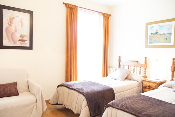 A hotel room with two beds, a wooden nightstand, a bedside table, and a window with curtains.