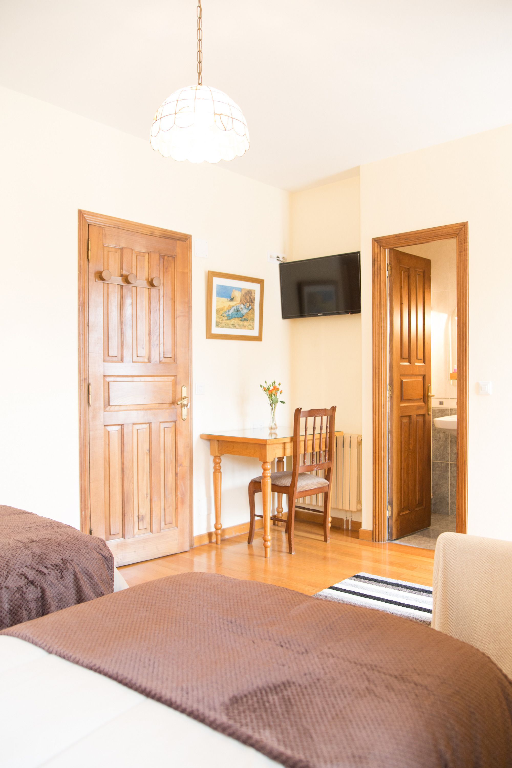 A room with a wooden door, a television mounted on the wall, a small table with a chair, and a bed with a brown blanket.