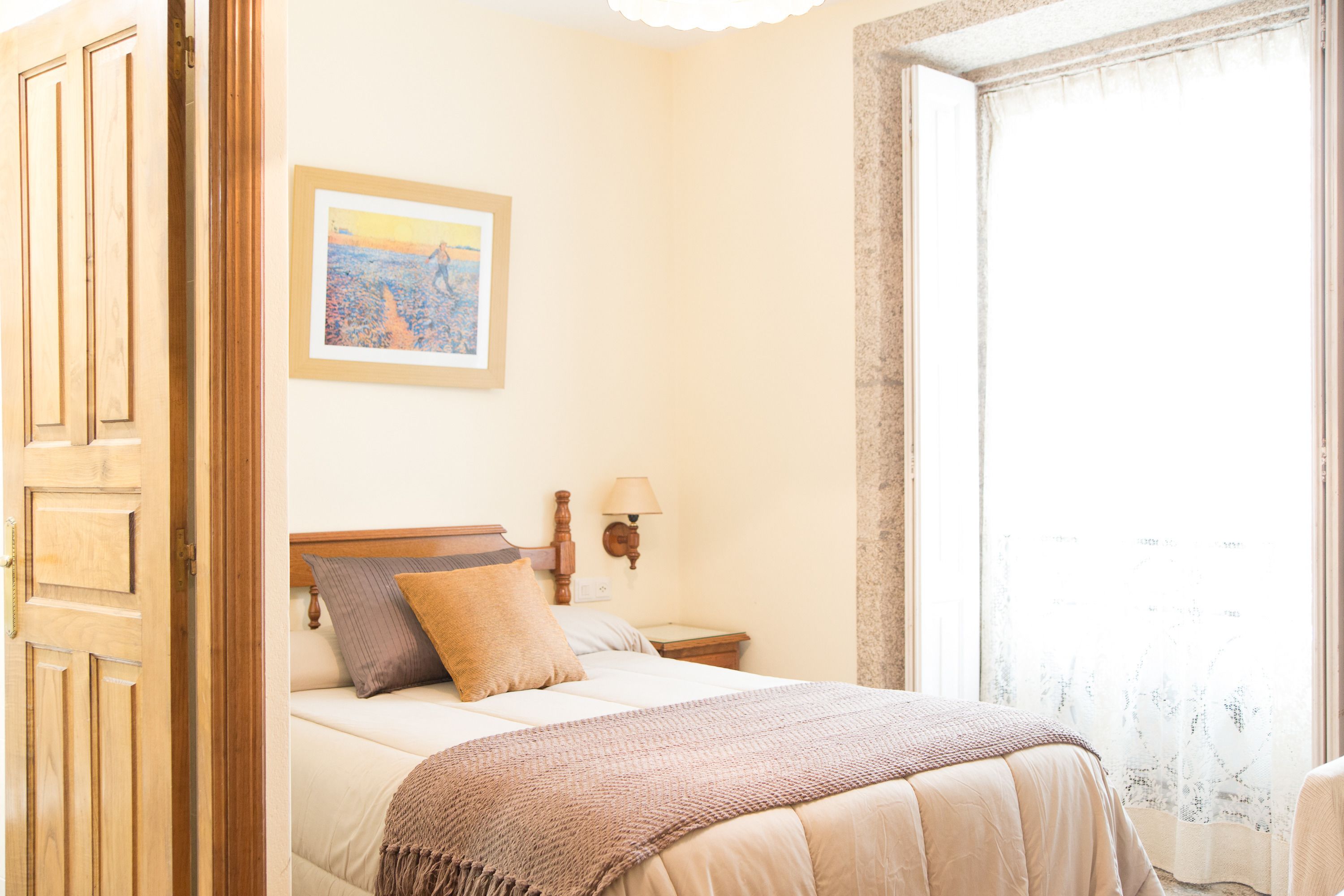 Een slaapkamer met een bed, een nachtkastje, een lamp, een lijst met een foto aan de muur en een raam met gordijnen.