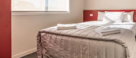 A neatly made bed with a red headboard, a window with blinds, and a carpeted floor.