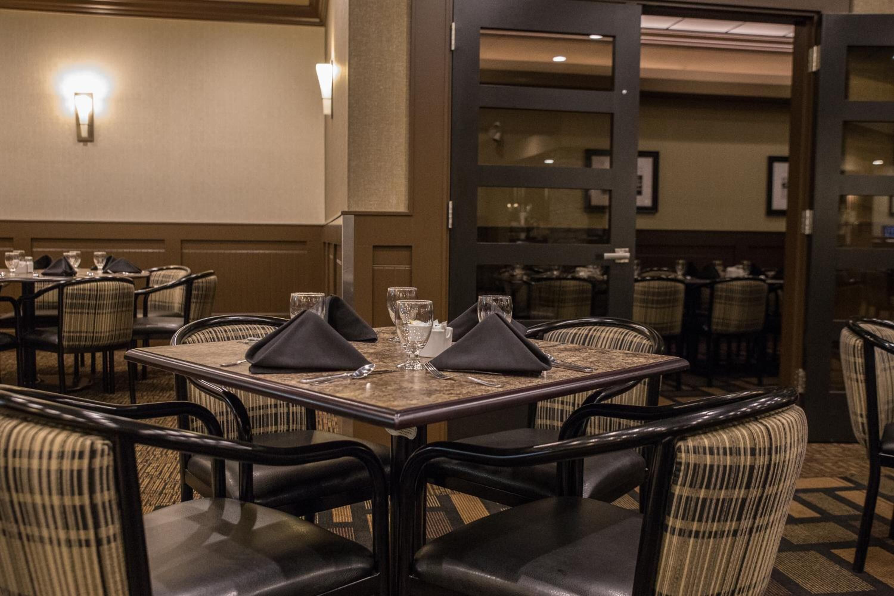A hotel dining area with tables set for four, each with a black napkin, a glass, and cutlery. The chairs are upholstered in a checkered pattern.