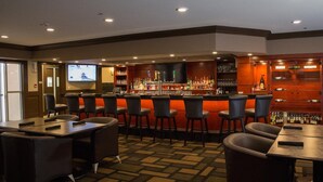 A bar area with multiple seating options, a television screen, and a variety of bottles and glasses on shelves.