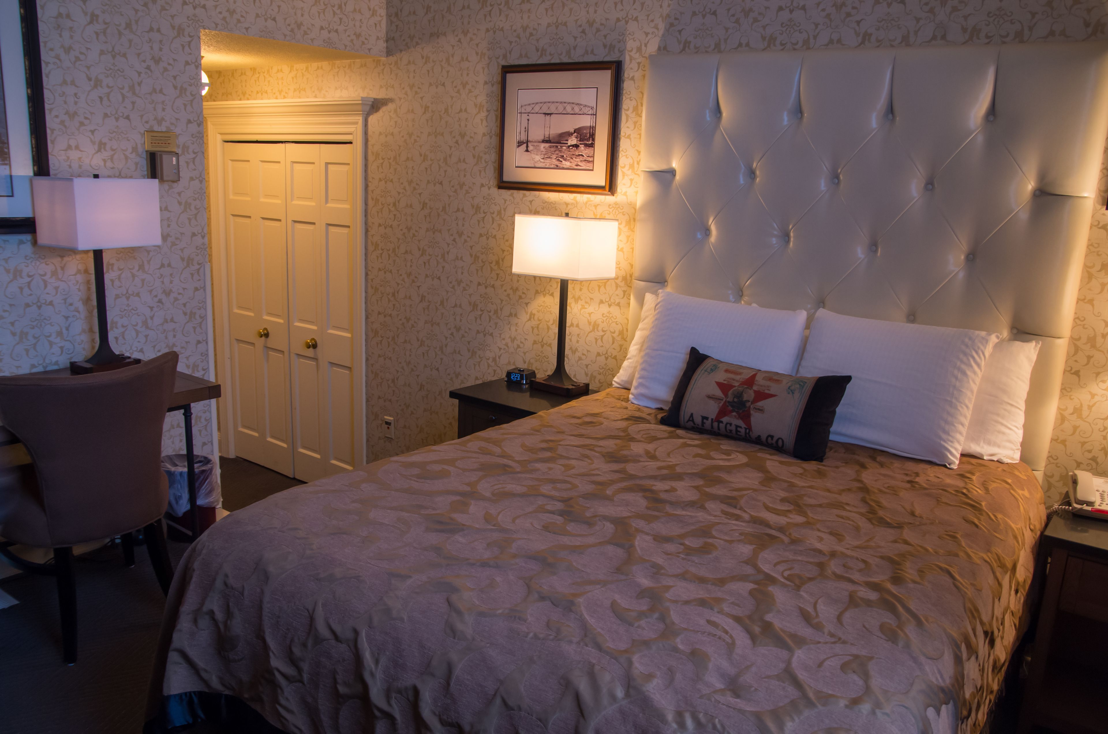 A hotel room with a large bed, a chair, a desk, a lamp, a framed picture on the wall, and a door.