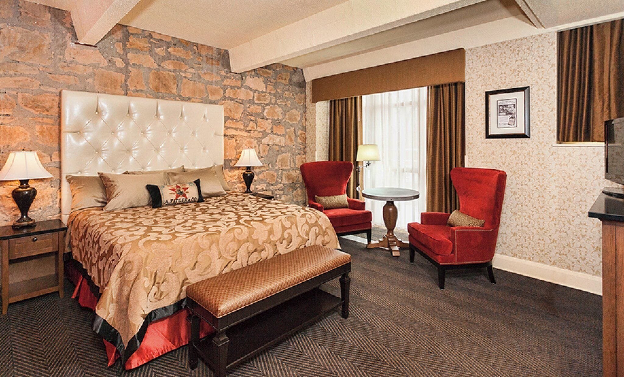 A hotel room with a large bed, a bench, two red armchairs, a small table, and a stone accent wall.