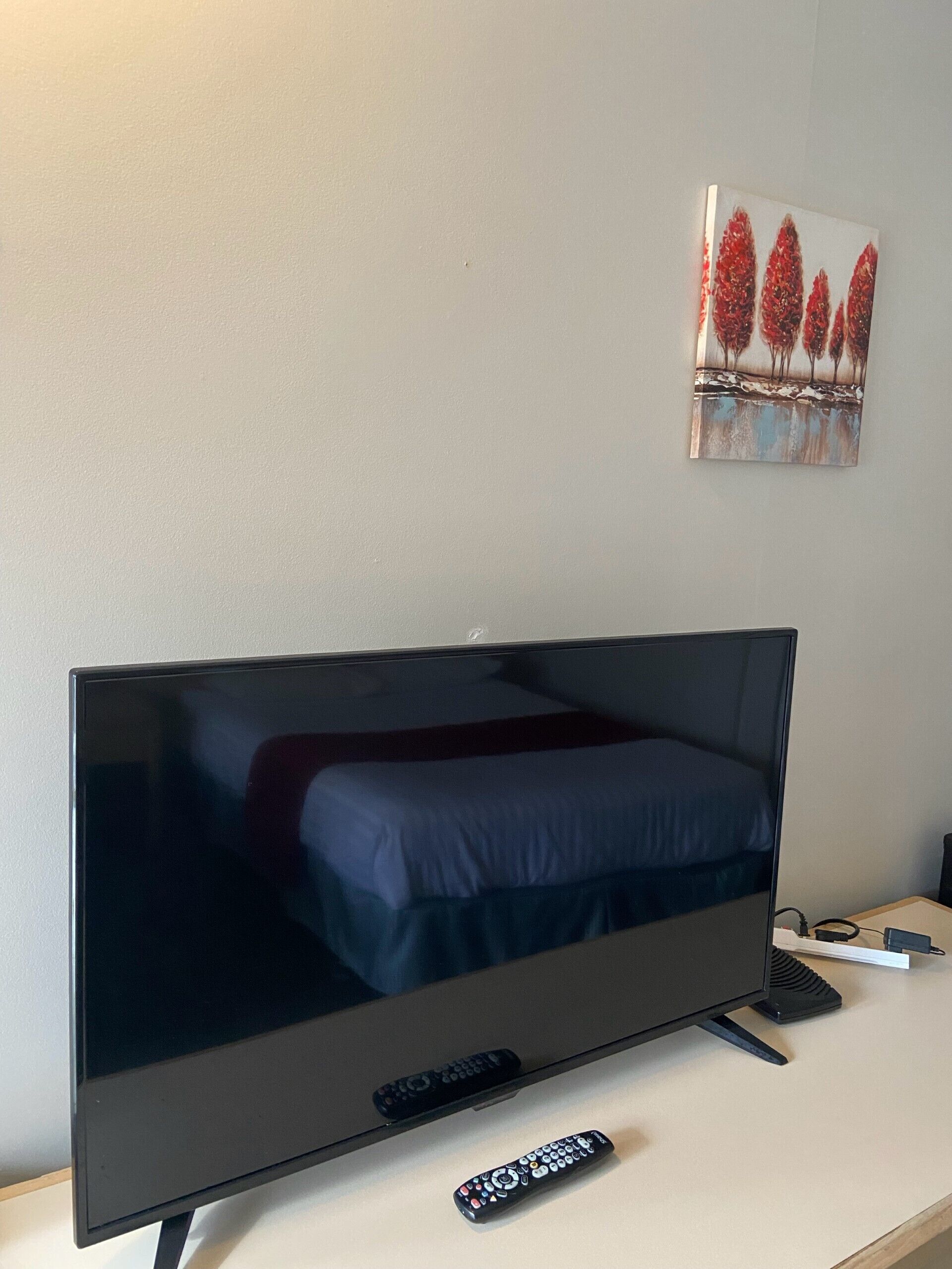 A hotel room with a large flat-screen TV, a bed with dark bedding, a remote control, and a small table with a phone and charger.