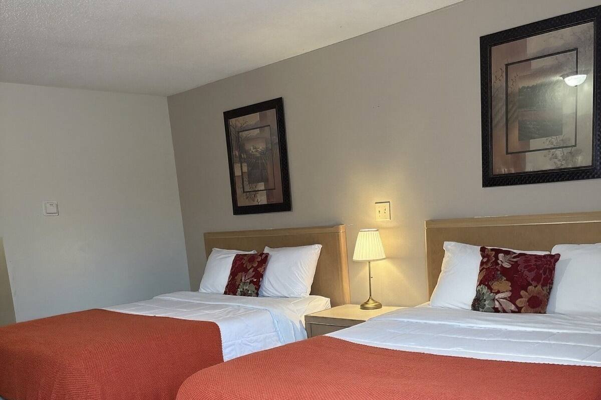A hotel room with two beds, each with a red blanket and white pillows, a nightstand with a lamp, and a framed picture on the wall.