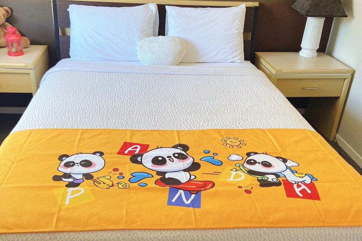 A bed with a white duvet, a yellow blanket with cartoon pandas and letters, a teddy bear, and a red lantern on a bedside table.