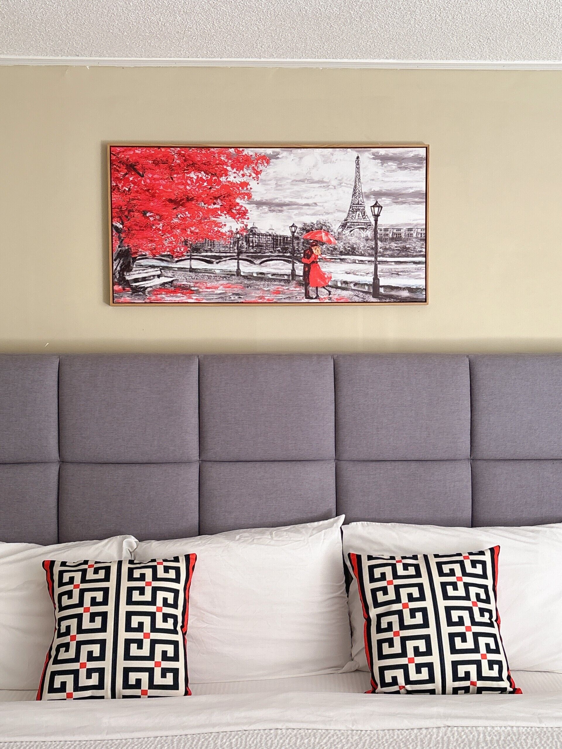 A room with a grey upholstered headboard, two patterned pillows, and a framed artwork depicting a red tree and the Eiffel Tower.