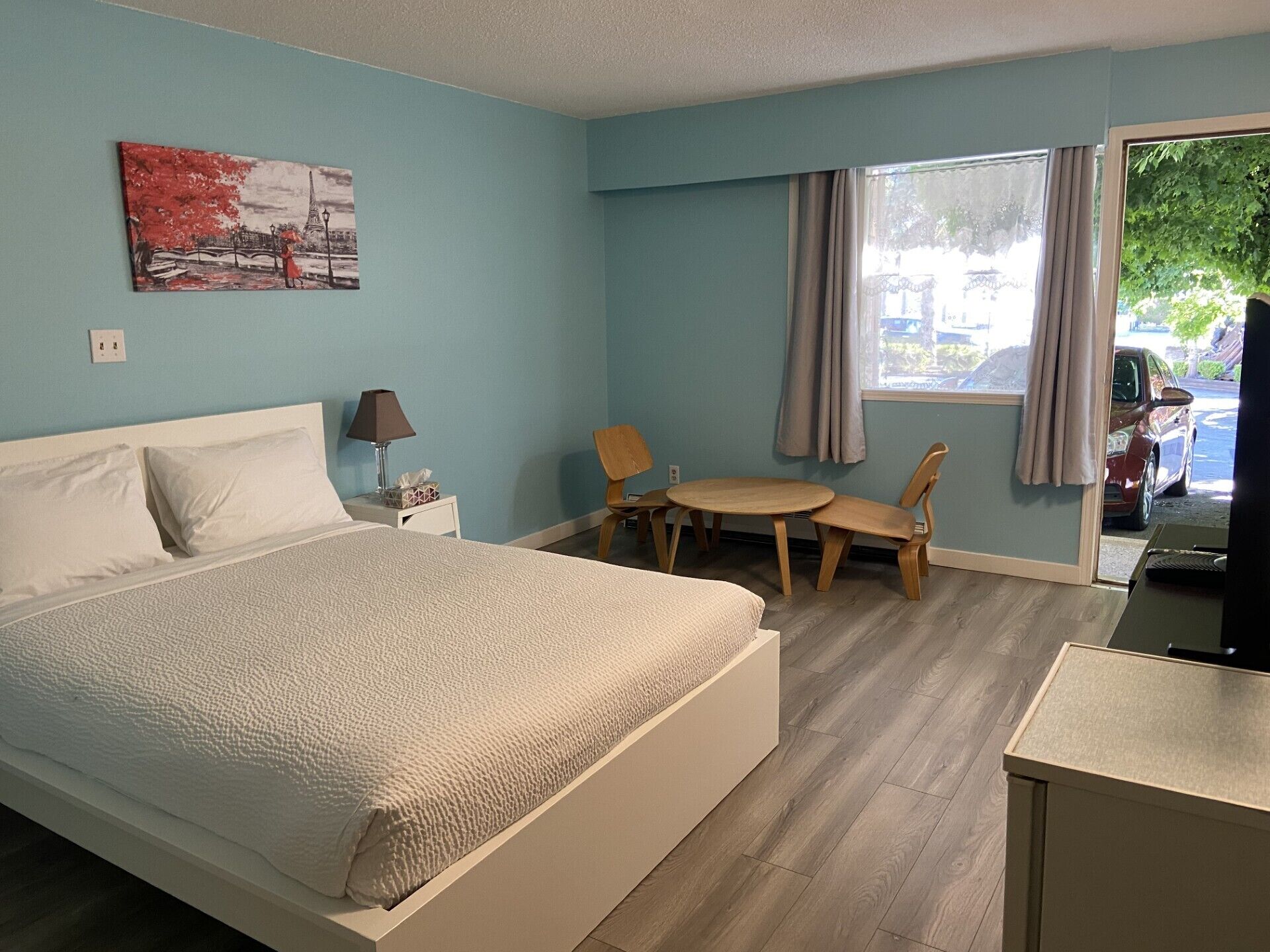 Habitación de hotel con cama, escritorio, silla, mesa, lámpara, un cuadro y una ventana con cortinas.