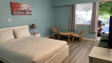 Habitación de hotel con cama, escritorio, silla, mesa, lámpara, un cuadro y una ventana con cortinas.