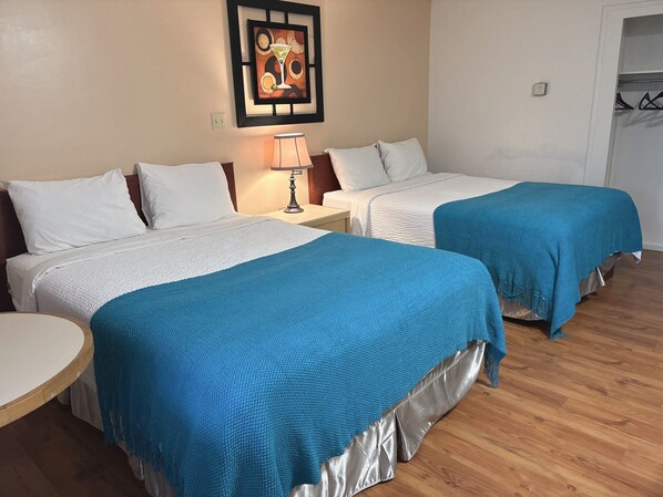 Habitación de hotel con dos camas, cada una con ropa blanca y una manta azul. Hay una mesita con una lámpara, y un cuadro en la pared.
