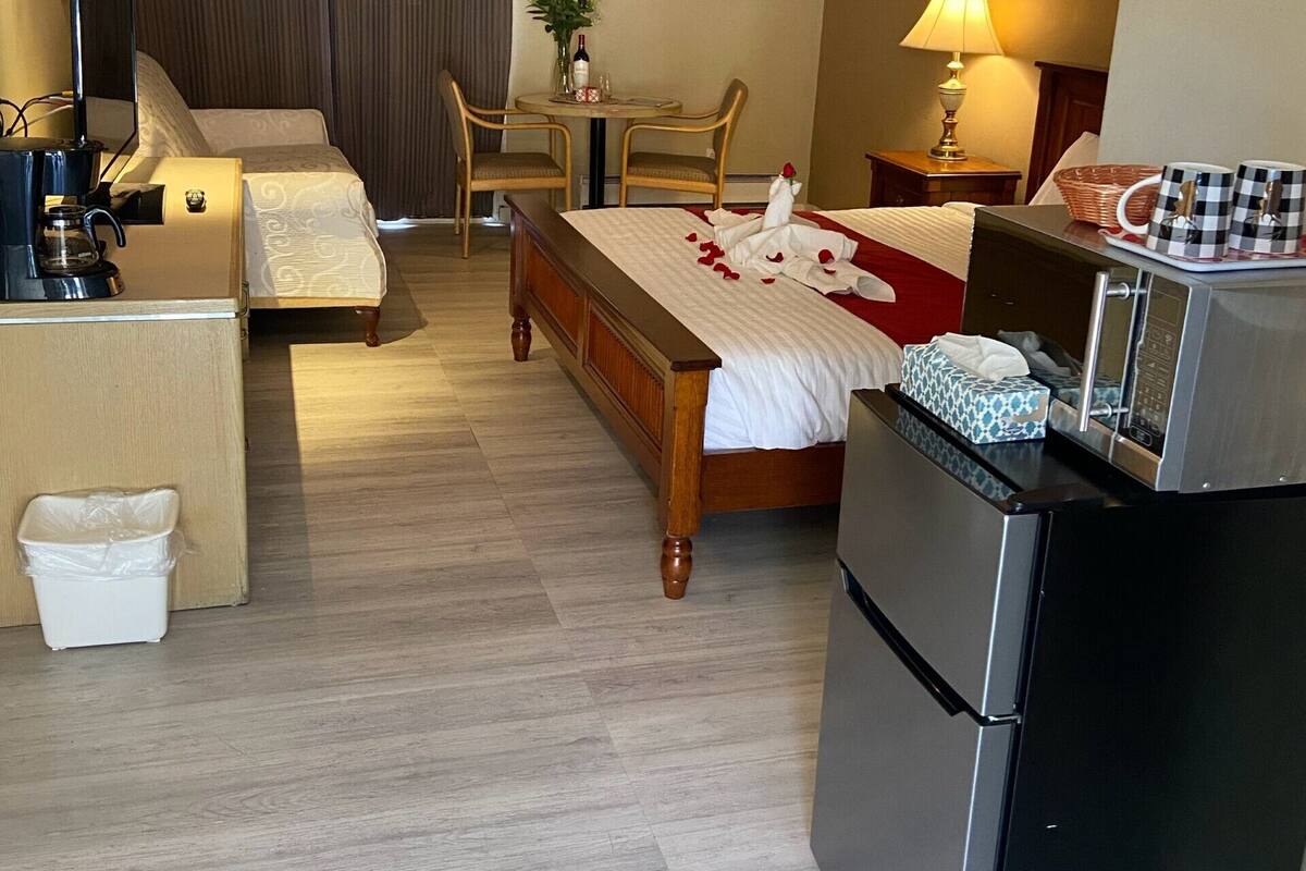 A hotel room with a bed, a desk, a refrigerator, a coffee maker, a table with a vase of flowers, and a chair.