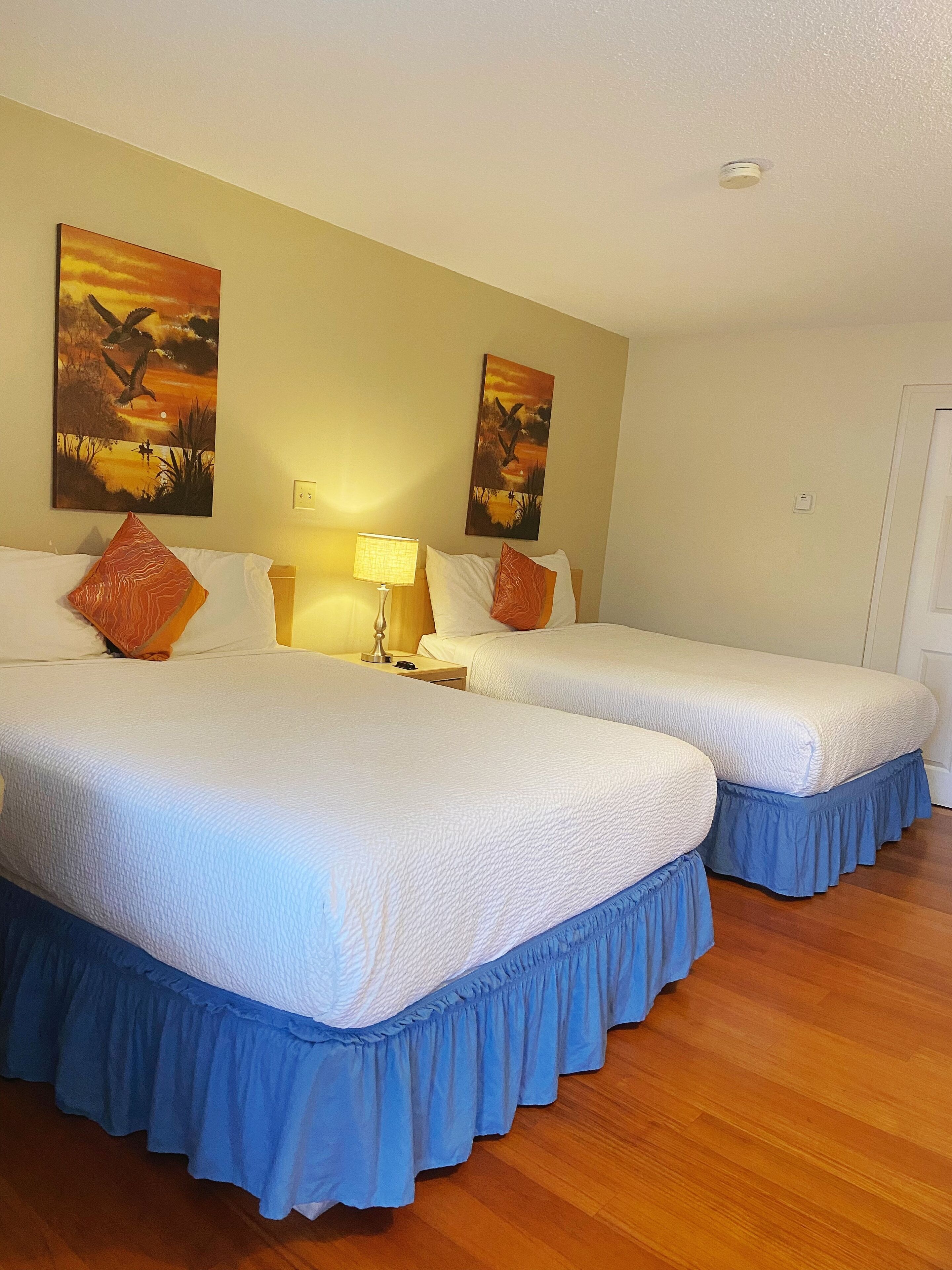 A hotel room with two beds, each with a blue skirted base, white bedding, and orange pillows. There are two wall-mounted paintings depicting birds in flight. A bedside lamp is on the left bed, and a power outlet is visible on the wall.