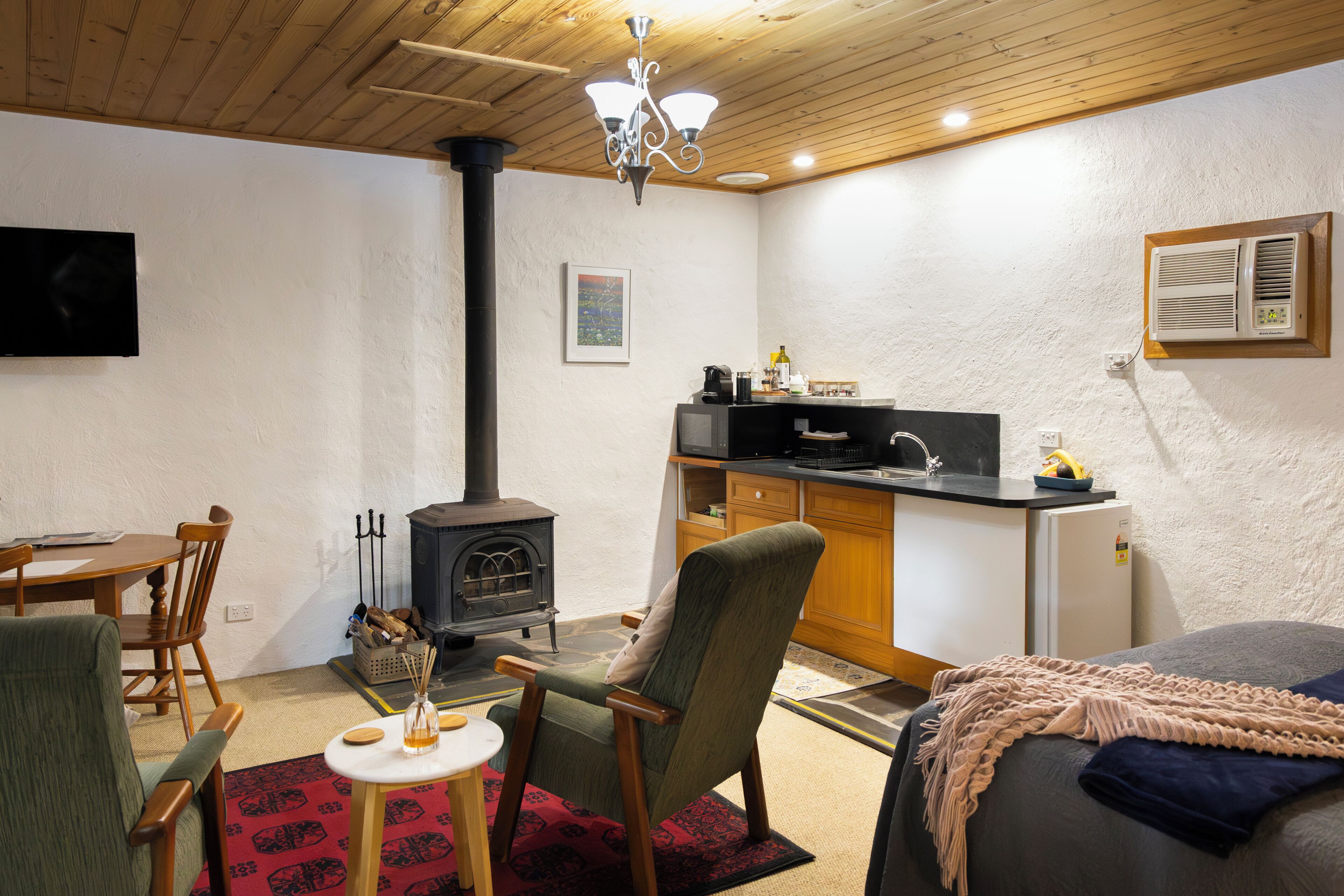 A cozy room with a wooden ceiling, a fireplace, a dining table, a kitchen area, and a bed.