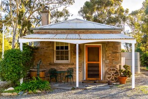 A stone cottage with a corrugated metal roof, a wooden door, and a small porch with two green metal chairs and a table. - Reillys Wines Heritage Cottages (Mintaro)