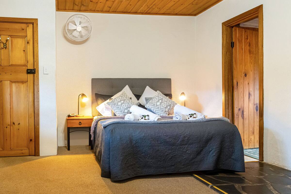 A bedroom with a bed, bedside table, lamp, and a wall-mounted fan.