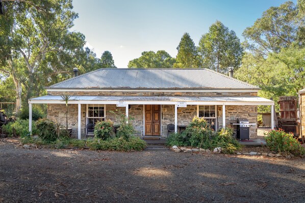 A stone house with a metal roof, a front porch, and a barbecue area. - Reillys Wines Heritage Cottages (Mintaro)