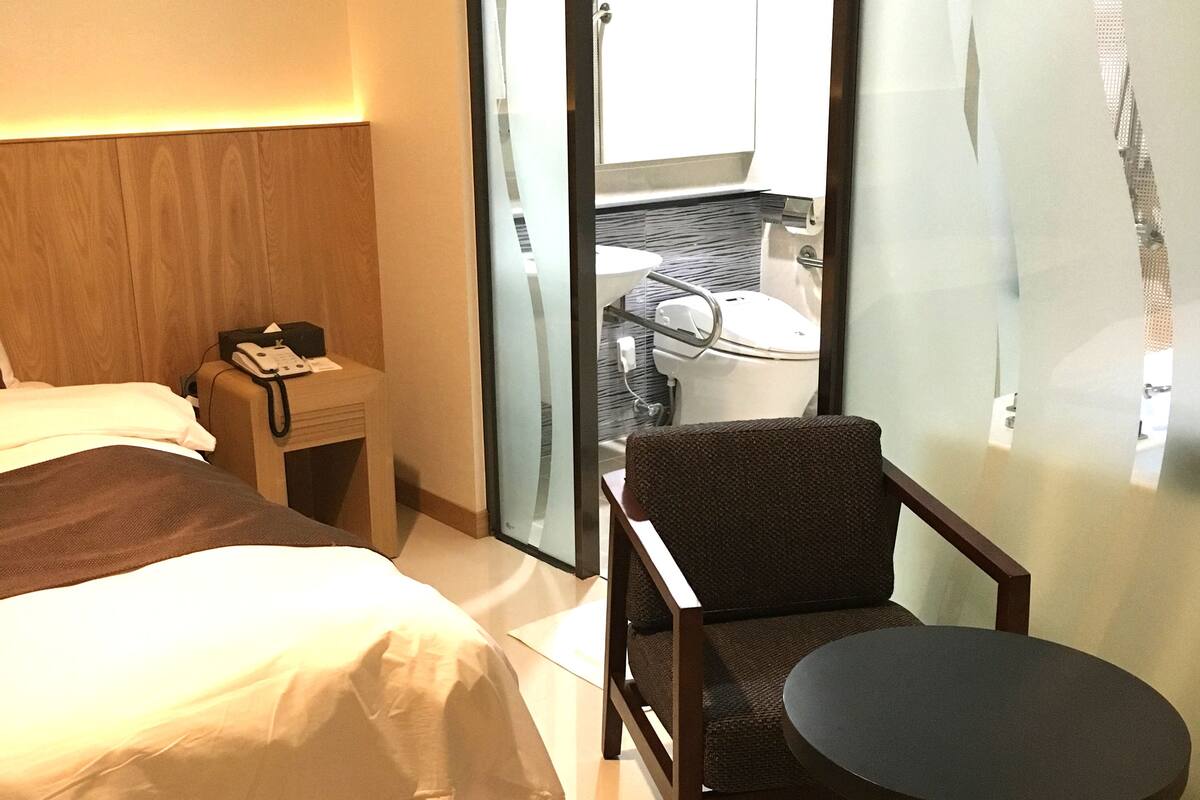 A hotel room with a bed, a small table, a chair, a nightstand, and a view of the bathroom through a glass door.