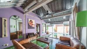 A room with a purple wall, wooden ceiling beams, a green tiled floor, a brown leather sofa, a wooden coffee table, and a green floor lamp. - Villa West (Seminyak)