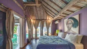 A bedroom with a wooden ceiling, a bed with a patterned bedspread, a painting on the wall, and a view of the outside. - Villa West (Seminyak)