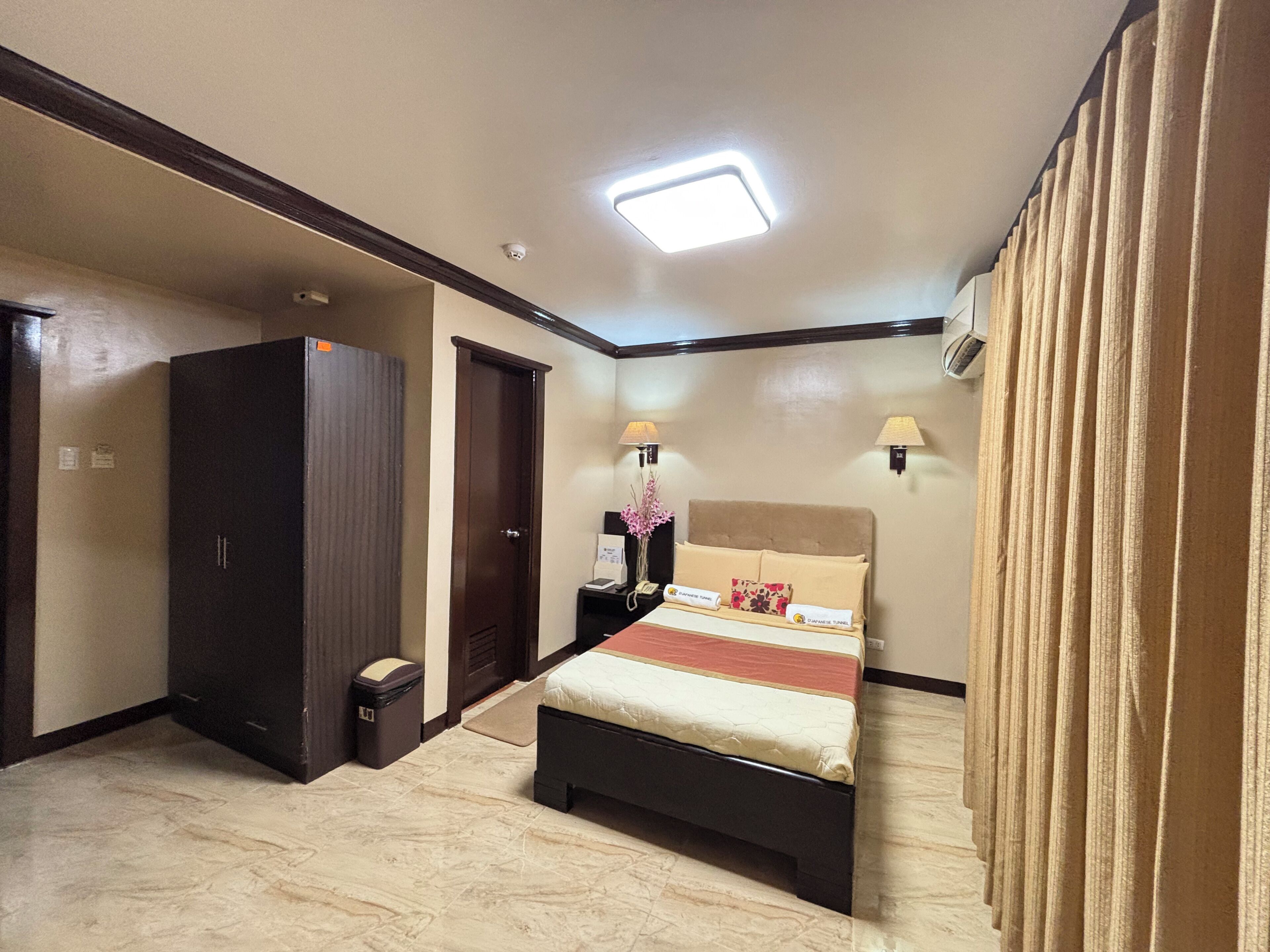 A hotel room with a bed, a wardrobe, a trash bin, a nightstand with a lamp, and a ceiling light.