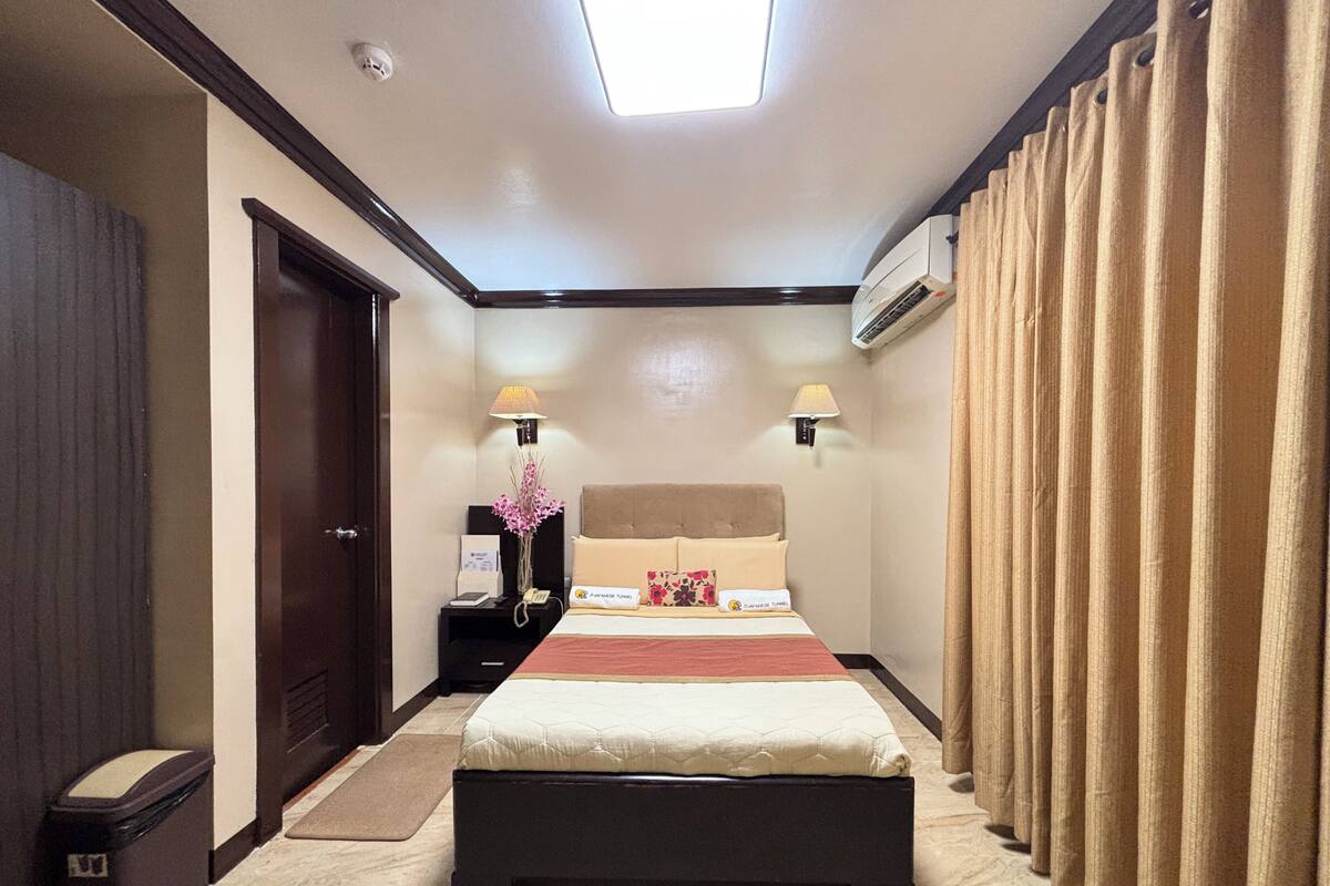 A hotel room with a bed, bedside lamps, an air conditioning unit, a nightstand, a trash bin, and a window with curtains.