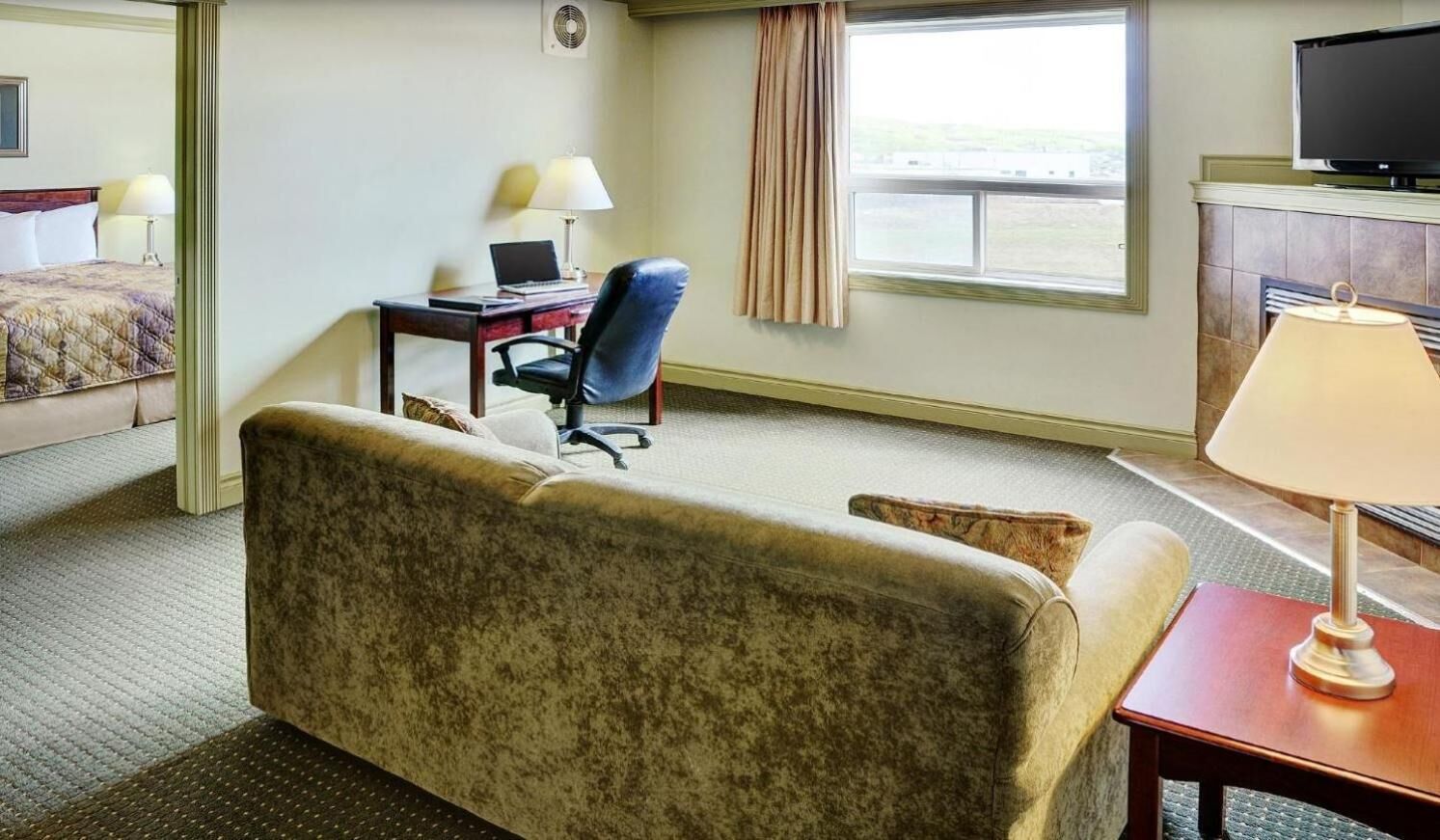 Habitación de hotel con cama, escritorio con una laptop, silla, televisor y ventana con cortinas.