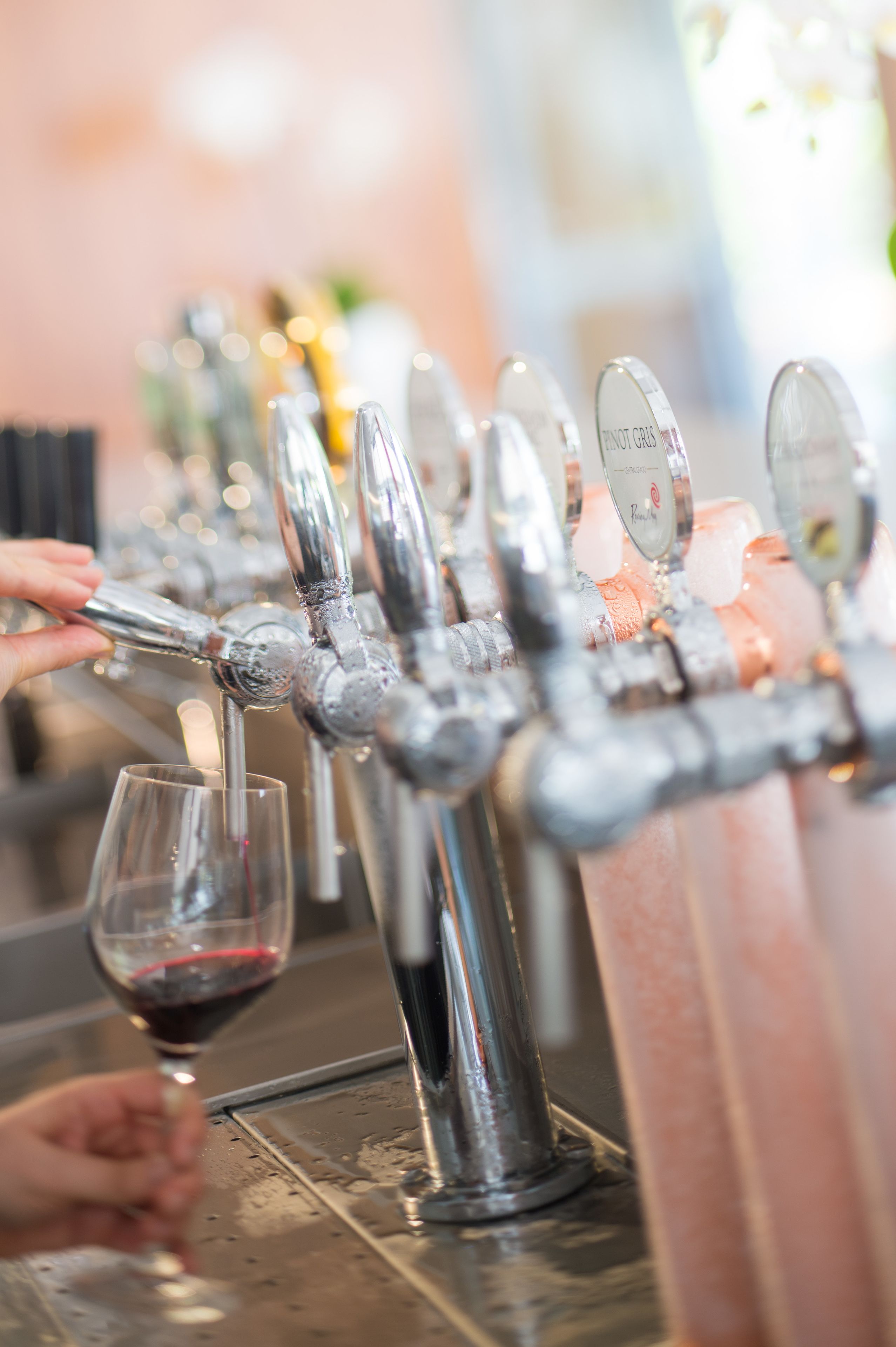 A bar with multiple taps, each with a label, and glasses of red wine on the counter.