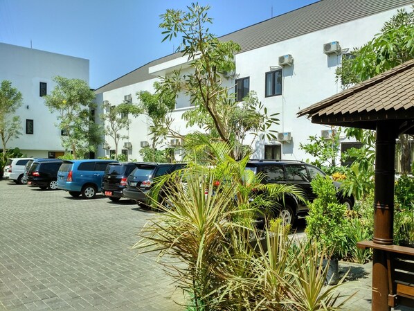 A parking area with several cars, a building with multiple windows and air conditioning units, and some greenery in the foreground.
